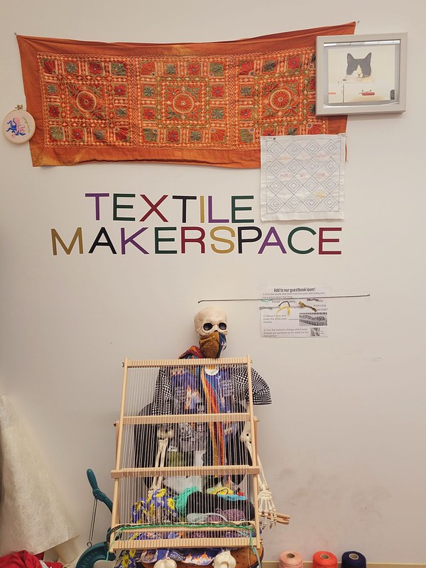 A skeleton holding the guestbook loom underneath a Textile Makerspace sign